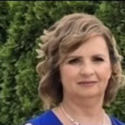 Portret kobiety o krótkich, kręconych blond włosach, ubranej w niebieską bluzkę, na tle zielonych krzewów.