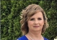 Portret kobiety o krótkich, kręconych blond włosach, ubranej w niebieską bluzkę, na tle zielonych krzewów.