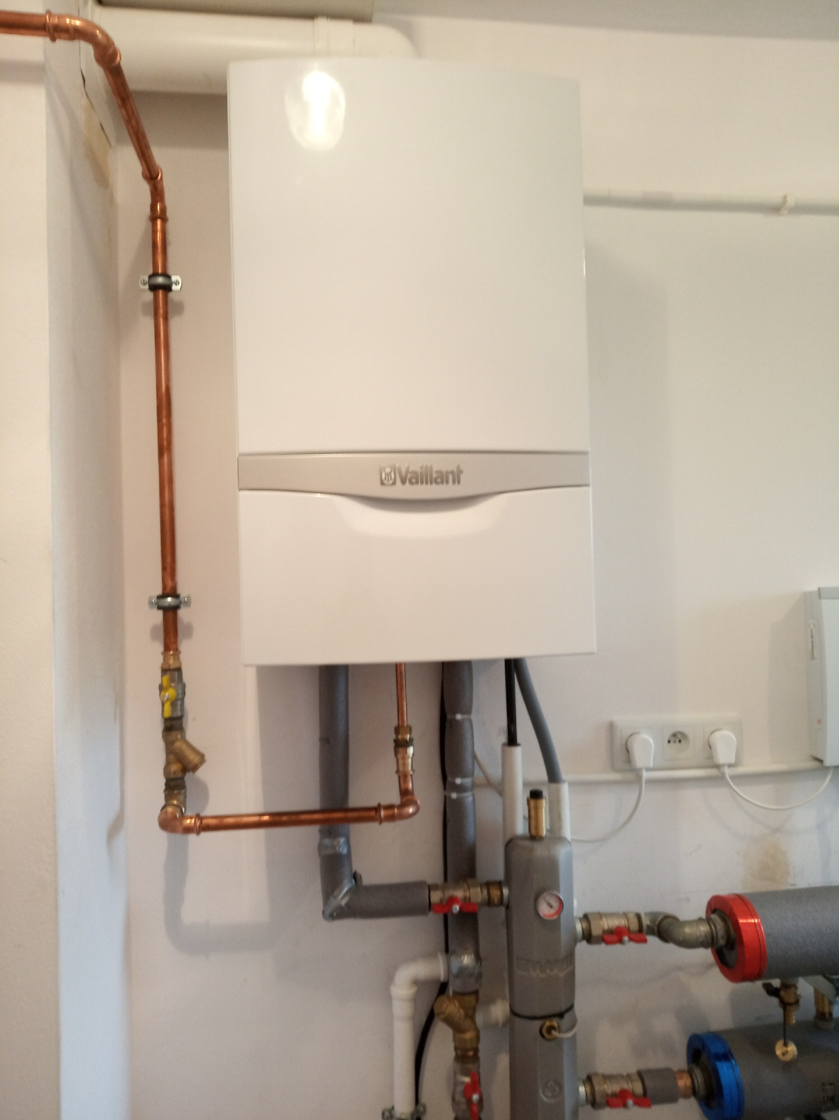 Biały, wiszący piec gazowy marki Vaillant z miedzianymi rurami i szarą armaturą, zamontowany na białej ścianie w pomieszczeniu technicznym.