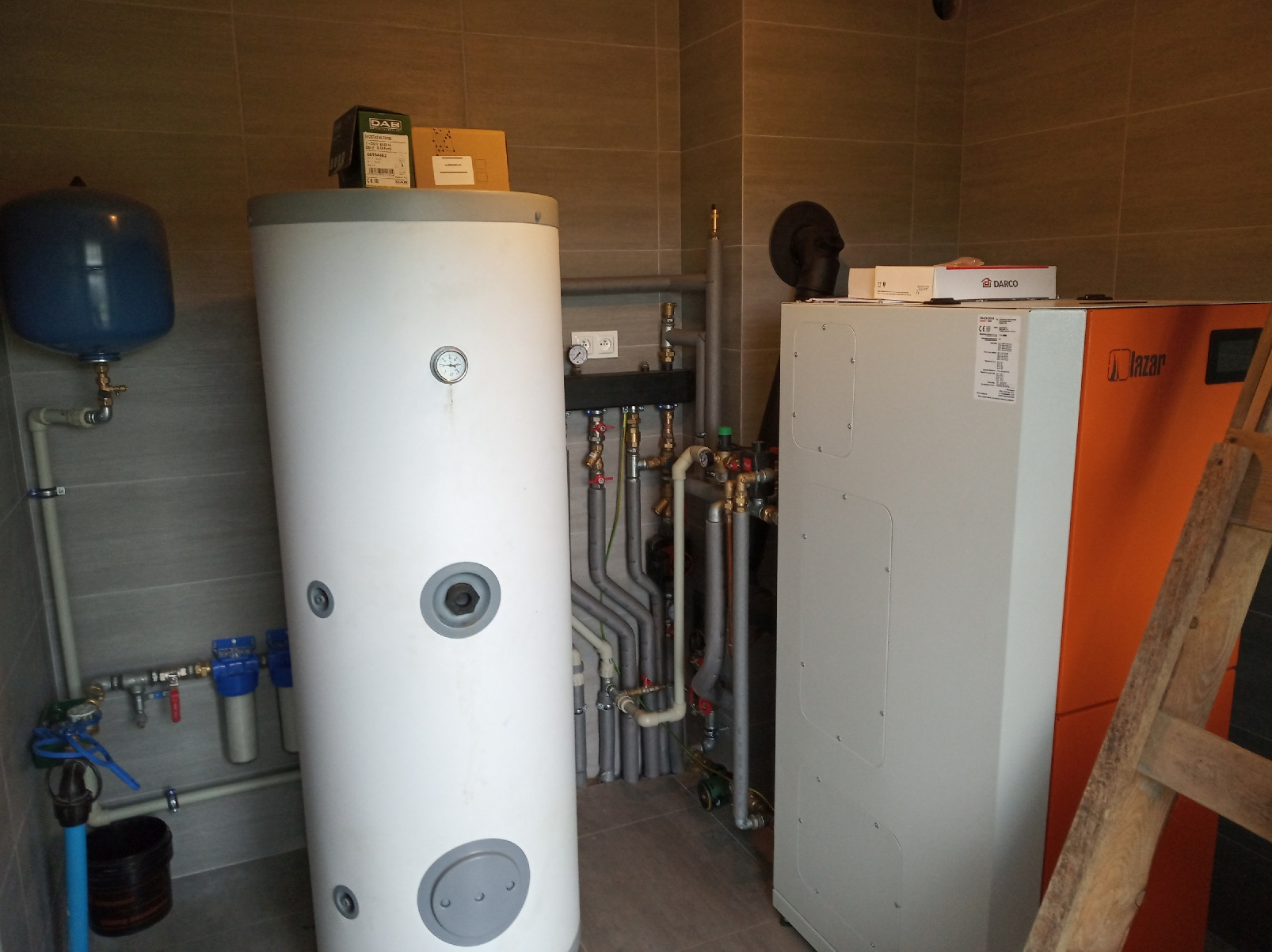 Kompaktowa kotłownia z białym zasobnikiem ciepłej wody, piecem z pomarańczowym panelem bocznym marki Lazar i rozbudowaną instalacją hydrauliczną z szarymi rurami w pomieszczeniu wyłożonym szarymi...