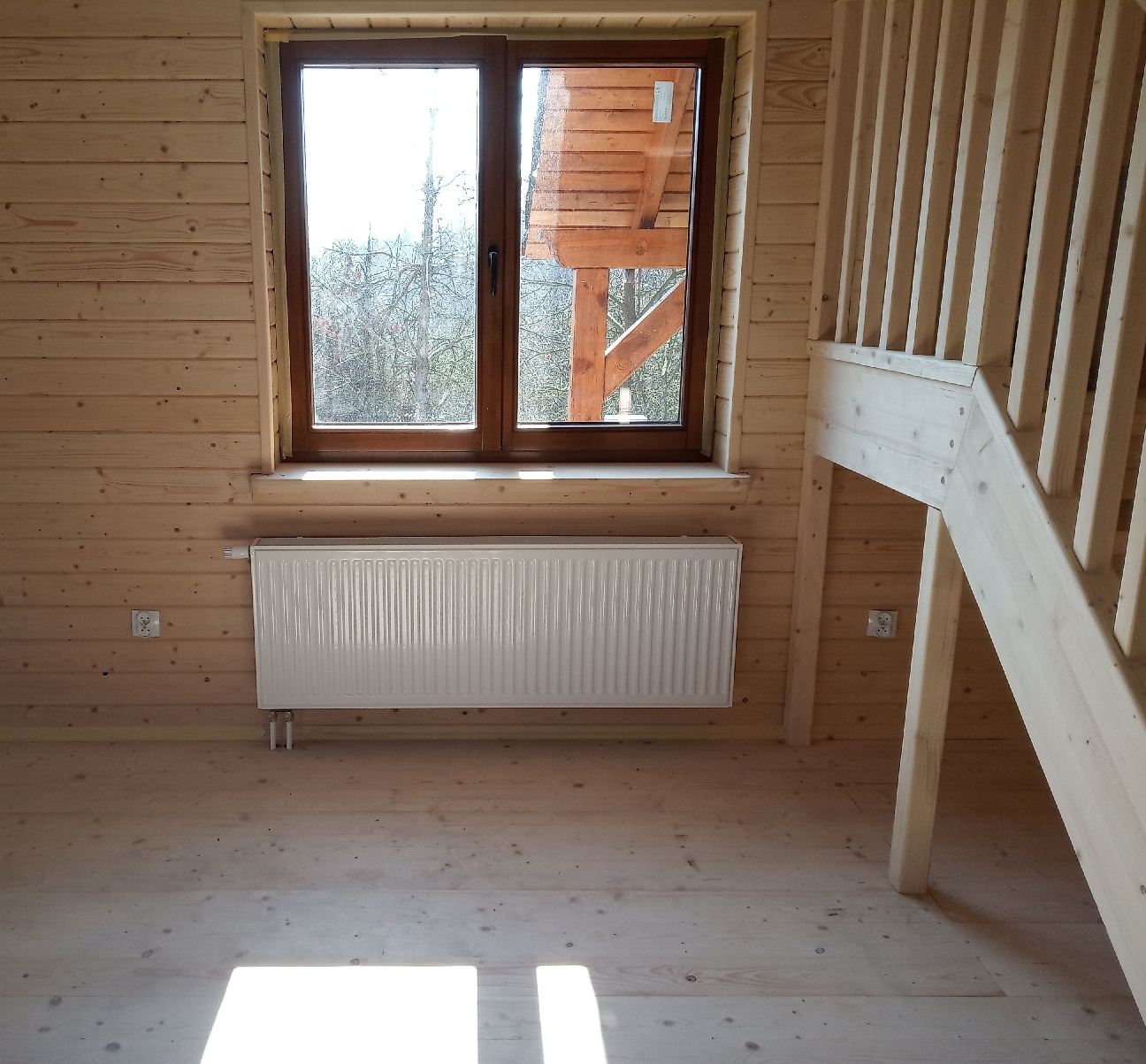 Biały grzejnik panelowy zamontowany pod oknem w drewnianym domu w Lublinie, widoczne drewniane ściany, podłoga i schody.