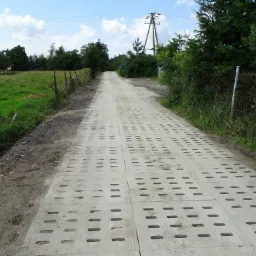 Długa droga gruntowa wzmocniona betonowymi płytami ażurowymi, prowadząca przez wiejski krajobraz z polami i drzewami, widoczna linia wysokiego napięcia.