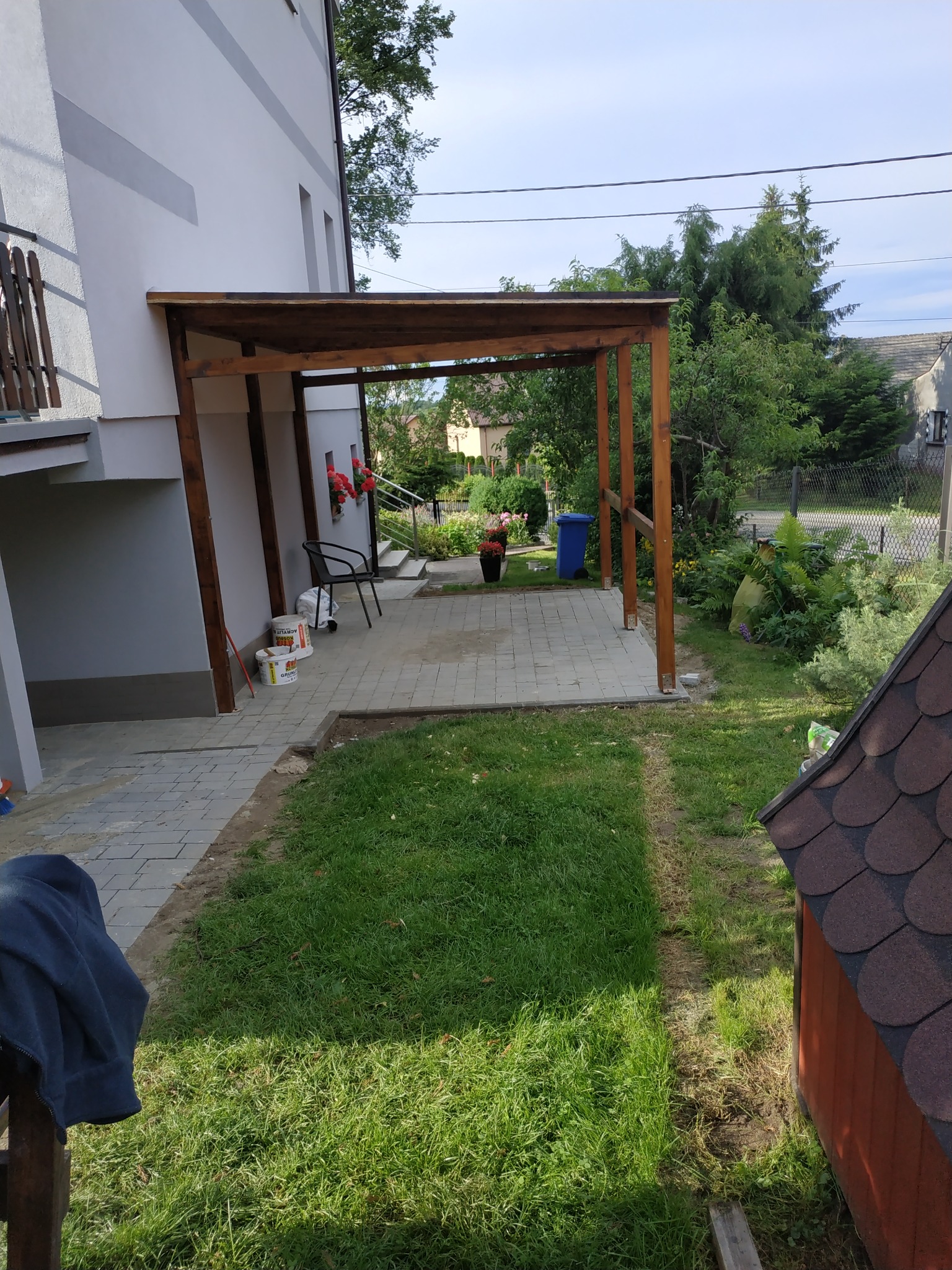 Nowo wybudowana drewniana pergola przy szarym domu z kostki brukowej, widoczne wiadra i pędzle, w tle ogród z kwiatami.
