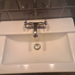 ART CLEAN Sp. z o.o. - Biała umywalka z chromowaną baterią i szarymi kafelkami w tle, widoczny fragment stopy i ubrania.