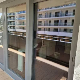 Przesuwne okno balkonowe z widoczną refleksją budynku naprzeciwko, na balkonie widoczne meble i wiadro.