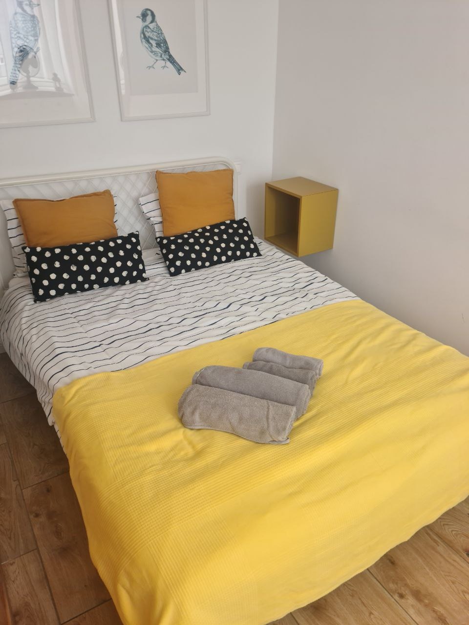 Ustawione ręczniki na żółtej kołdrze na łóżku w pokoju z drewnianą podłogą, obok biała ściana z dwoma obrazkami i żółtą szafką nocną.