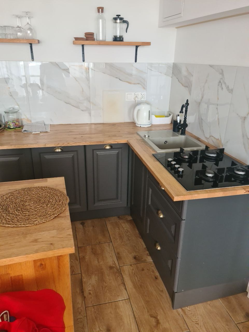 Czarny zestaw szafek kuchennych z drewnianym blatem, ceramiczna płyta grzewcza z czterema palnikami, zlewozmywak z czarną baterią, marmurowe płytki na ścianie, drewniane półki z dekoracjami.