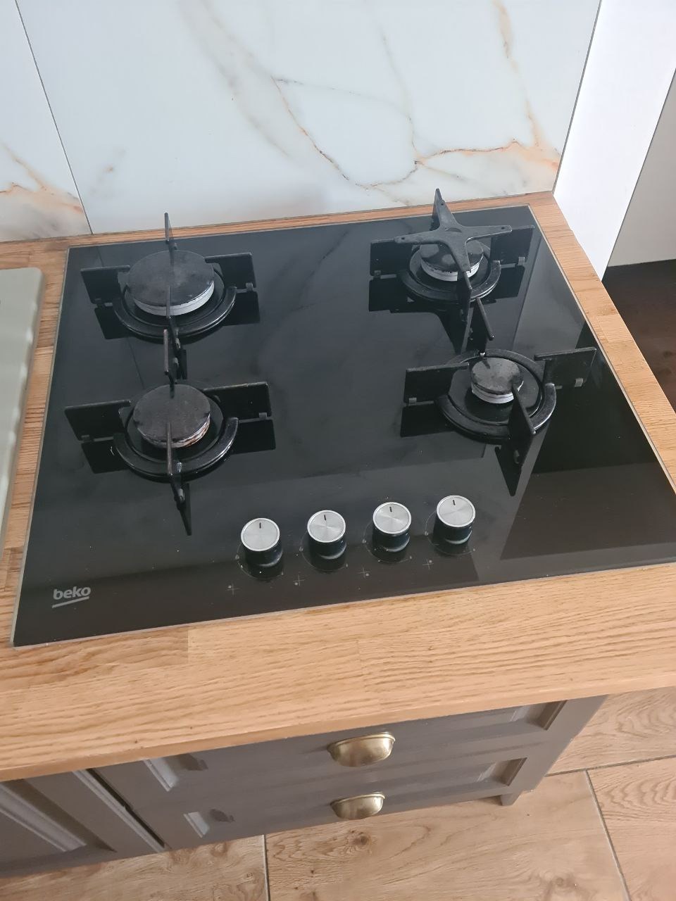 Czarna płyta gazowa Beko zamontowana na drewnianym blacie kuchennym, z białymi marmurkowymi płytkami w tle.