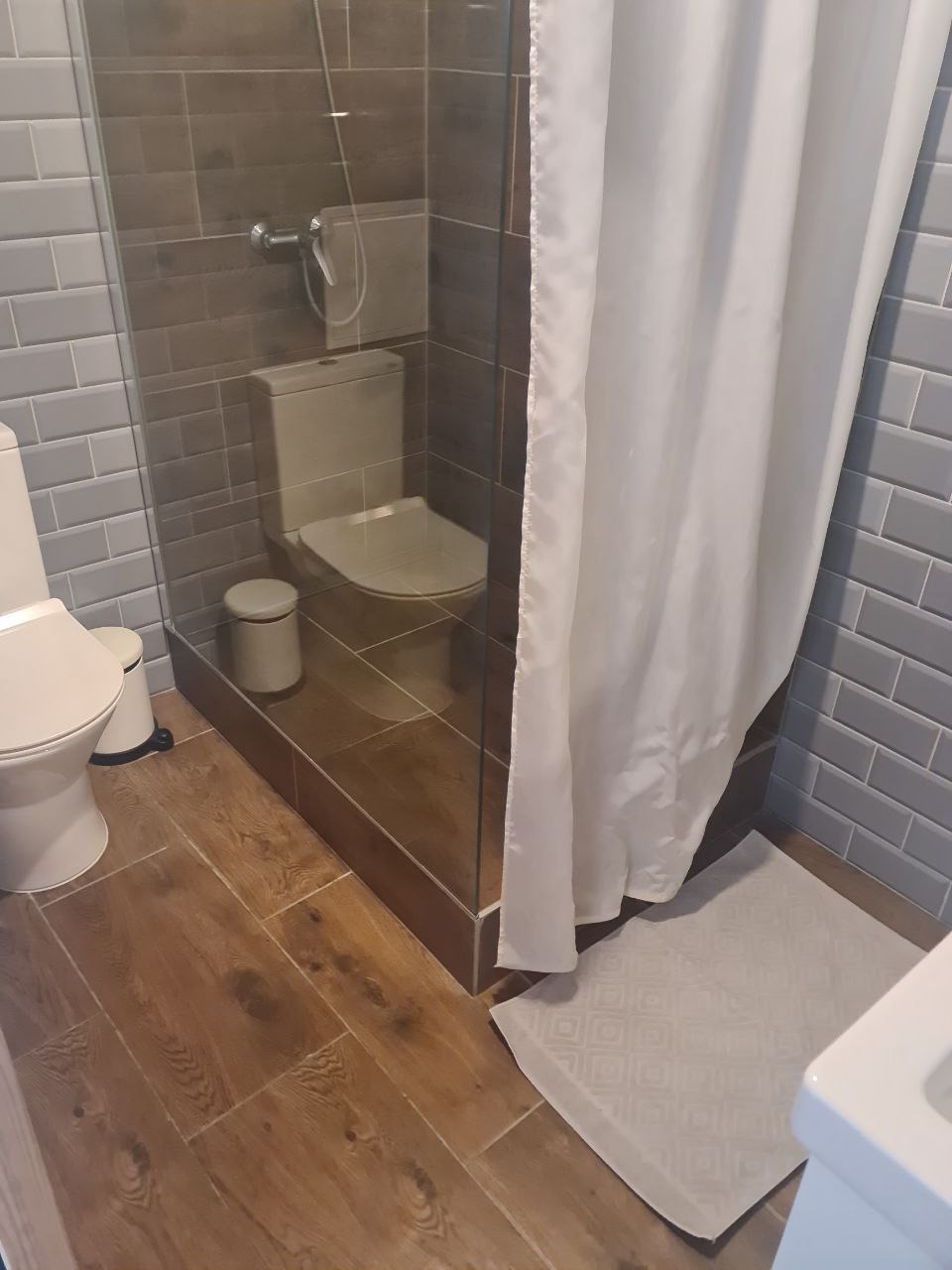 Umywalka i toaleta w łazience z kabiną prysznicową, brązowe płytki podłogowe i szare ścienne, biała zasłona prysznicowa i dywanik.