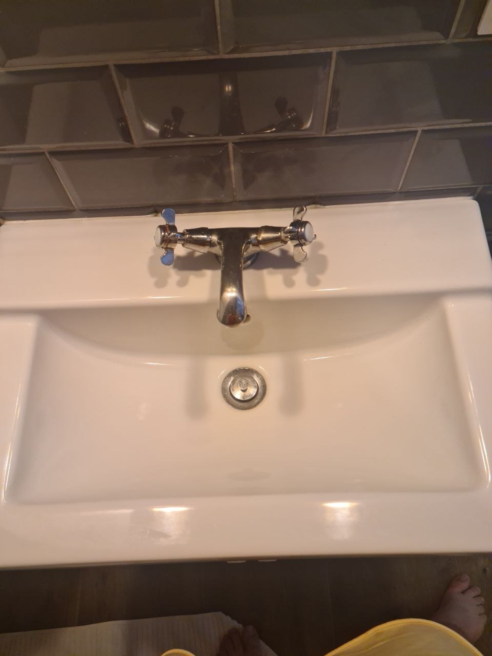 Biała umywalka z chromowaną baterią i szarymi kafelkami w tle, widoczny fragment stopy i ubrania.