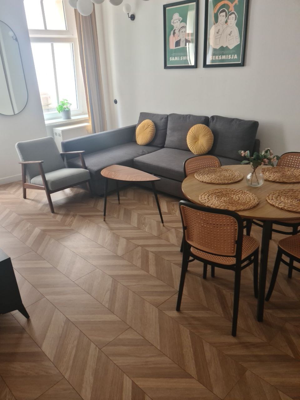 Jasne wnętrze mieszkania z szarą sofą, okrągłym stołem z plecionymi podkładkami, drewnianą podłogą ułożoną w jodełkę i plakatami na ścianie.