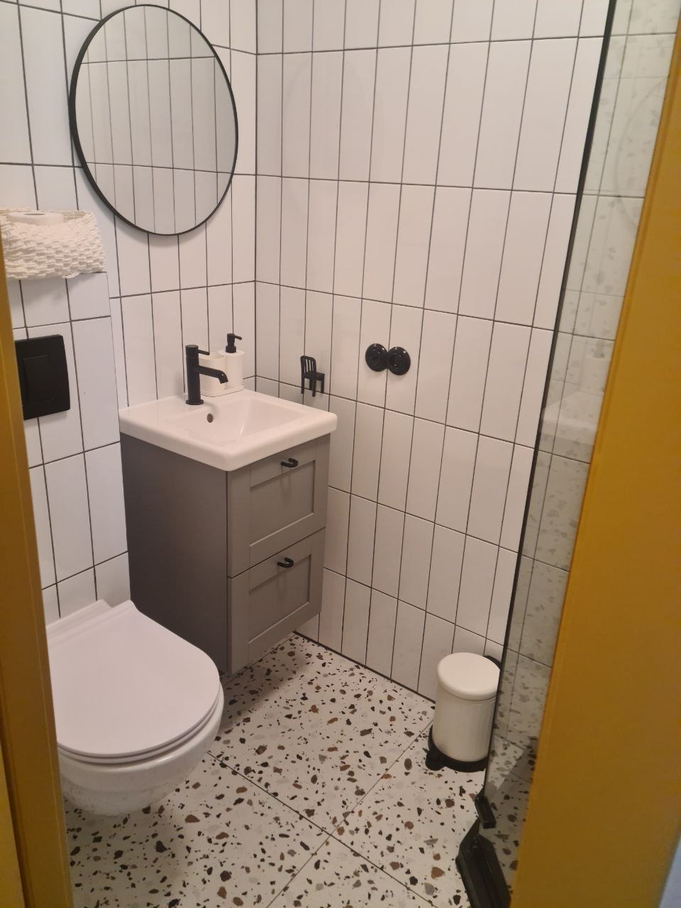 Wnętrze wyremontowanej łazienki z biało-szarą armaturą, mozaikową podłogą i okrągłym lustrem.