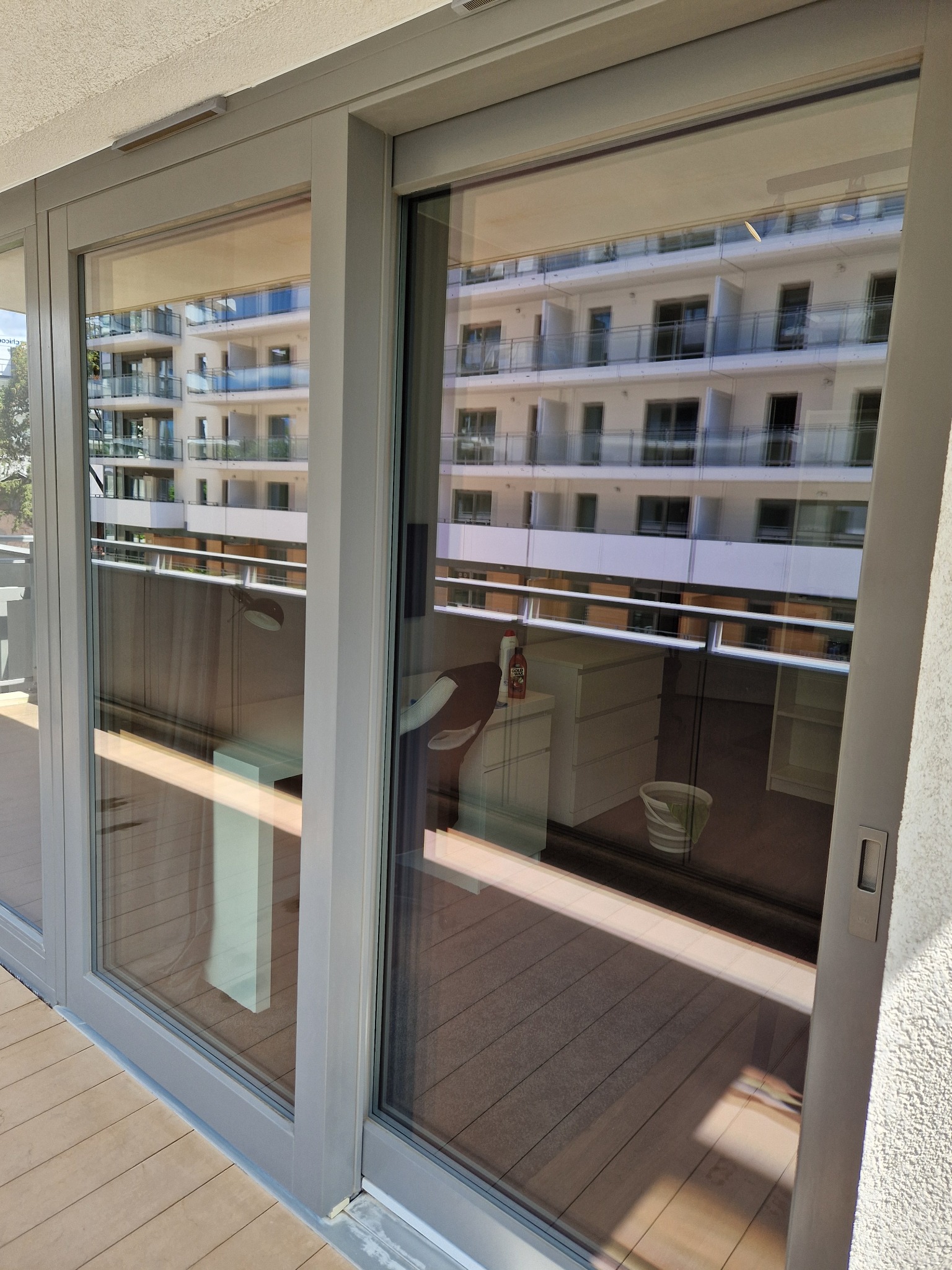 Przesuwne okno balkonowe z widoczną refleksją budynku naprzeciwko, na balkonie widoczne meble i wiadro.