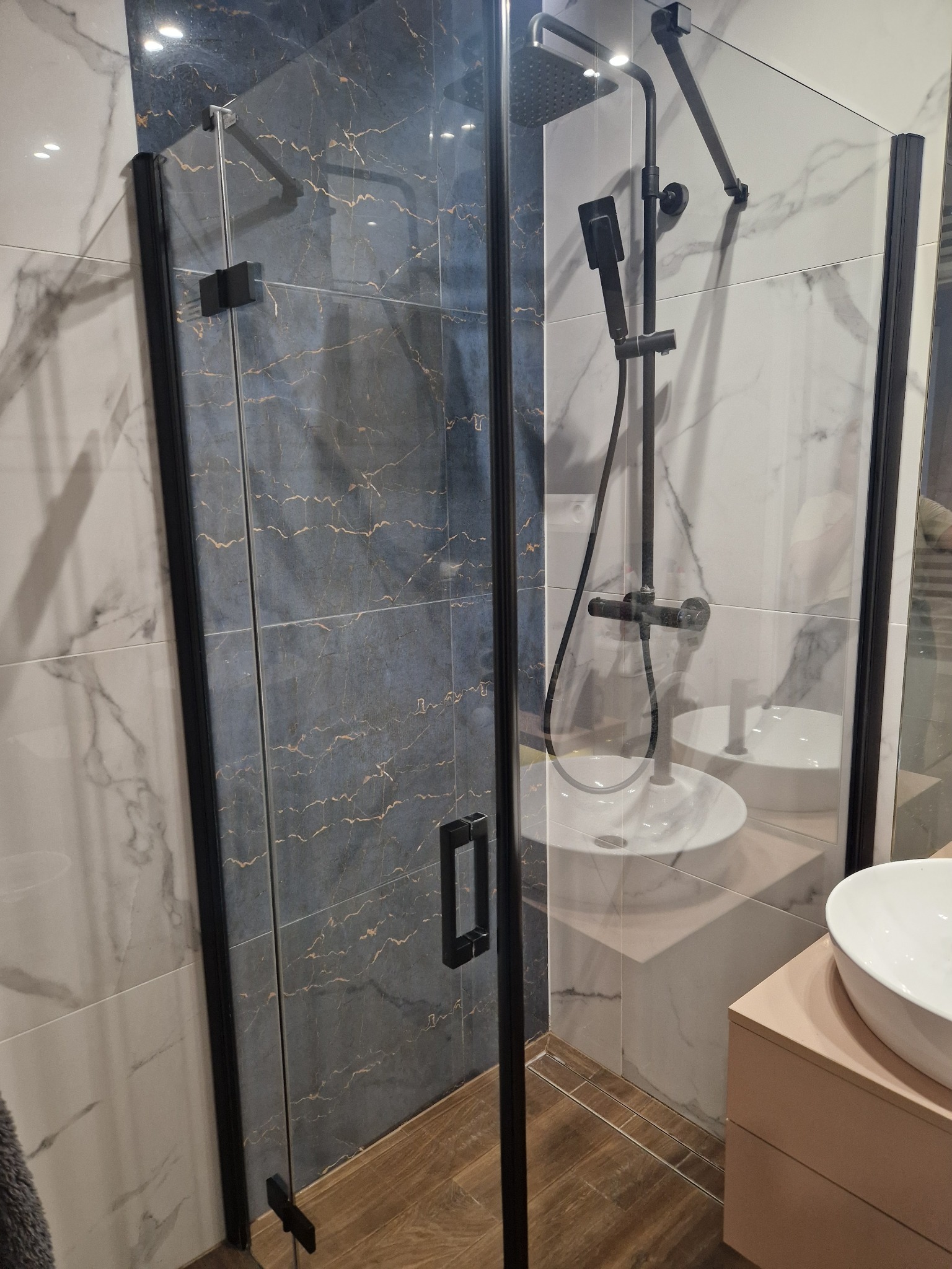 Szklana kabina prysznicowa z czarnymi profilami, widoczne marmurowe płytki w odcieniach szarości i bieli, oraz czarny zestaw prysznicowy z deszczownicą i słuchawką.