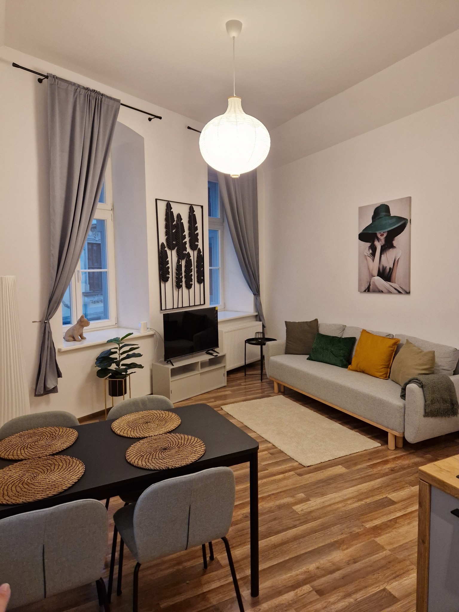 Jasne wnętrze salonu z szarymi zasłonami, drewnianą podłogą, szarą sofą z kolorowymi poduszkami, czarnym stolikiem kawowym i stołem z plecionymi podkładkami, oświetlone wiszącą lampą.