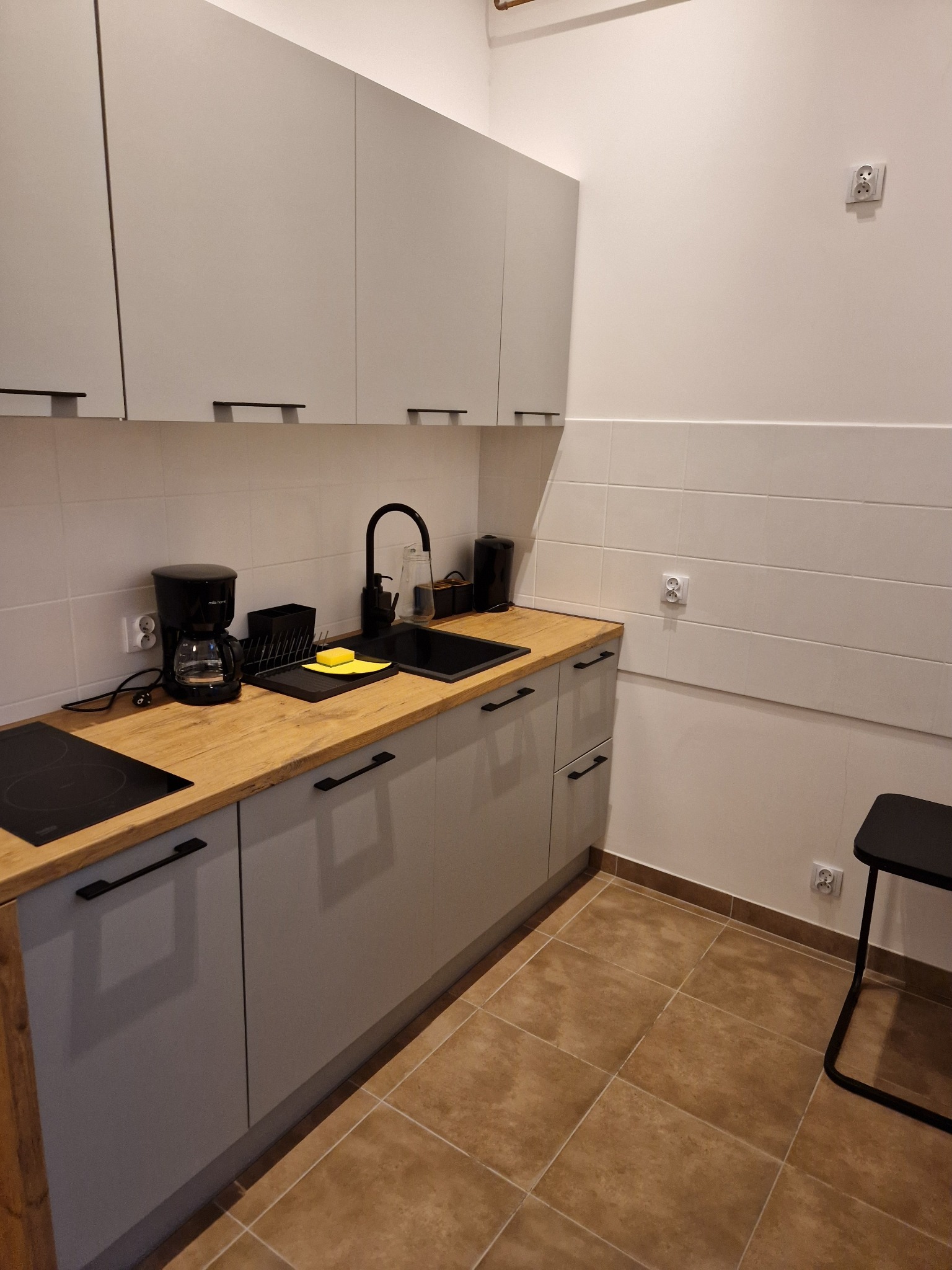 Uporządkowany aneks kuchenny z szarymi szafkami, drewnianym blatem, czarnym zlewem i baterią, ekspres do kawy oraz żółta gąbka do naczyń na suszarce.