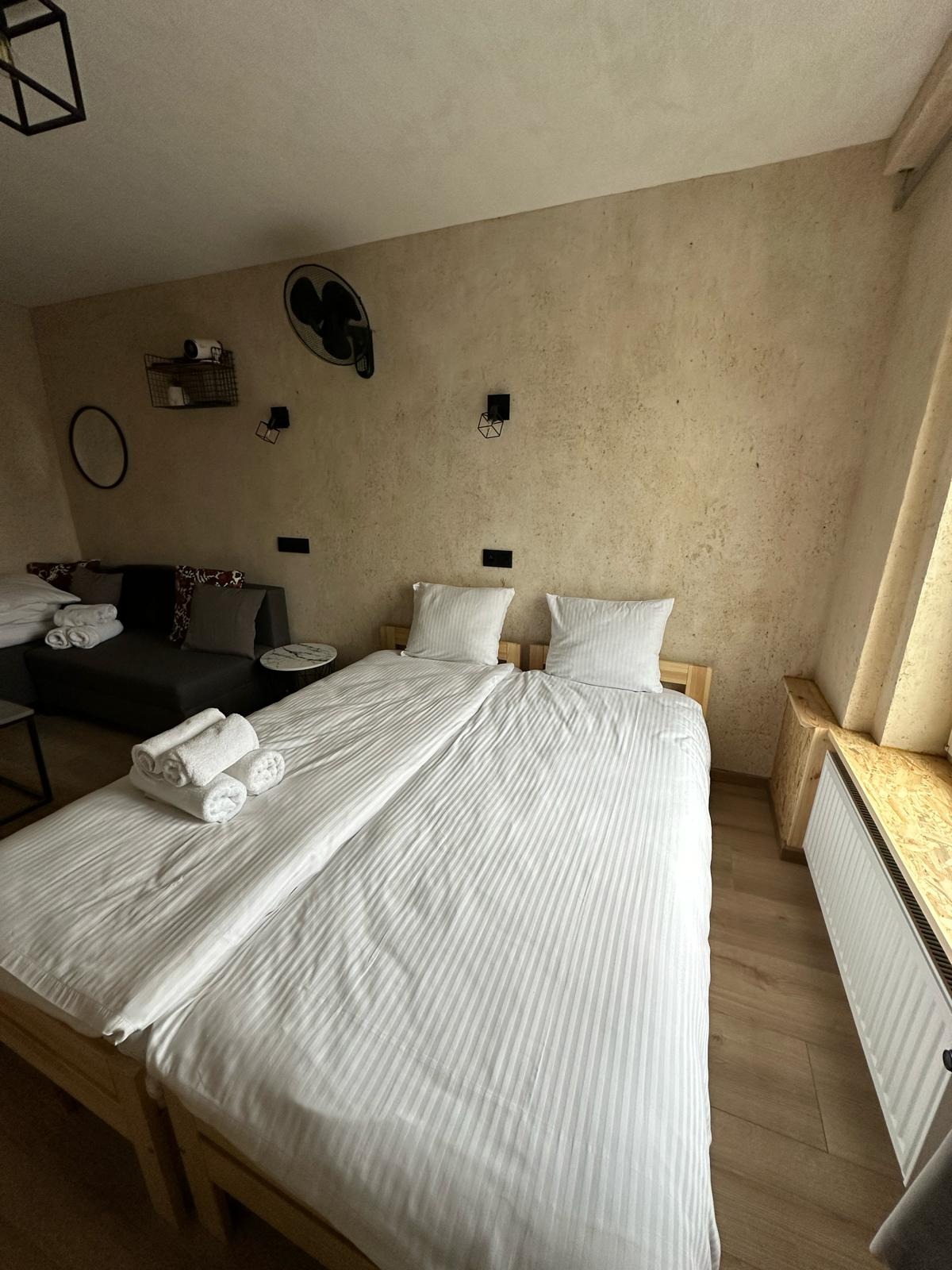 Pokój hotelowy z dwoma pojedynczymi łóżkami z białą pościelą w paski, złożone białe ręczniki na łóżku, sofa z poduszkami w tle, wentylator i lampy na ścianie o szorstkiej fakturze.