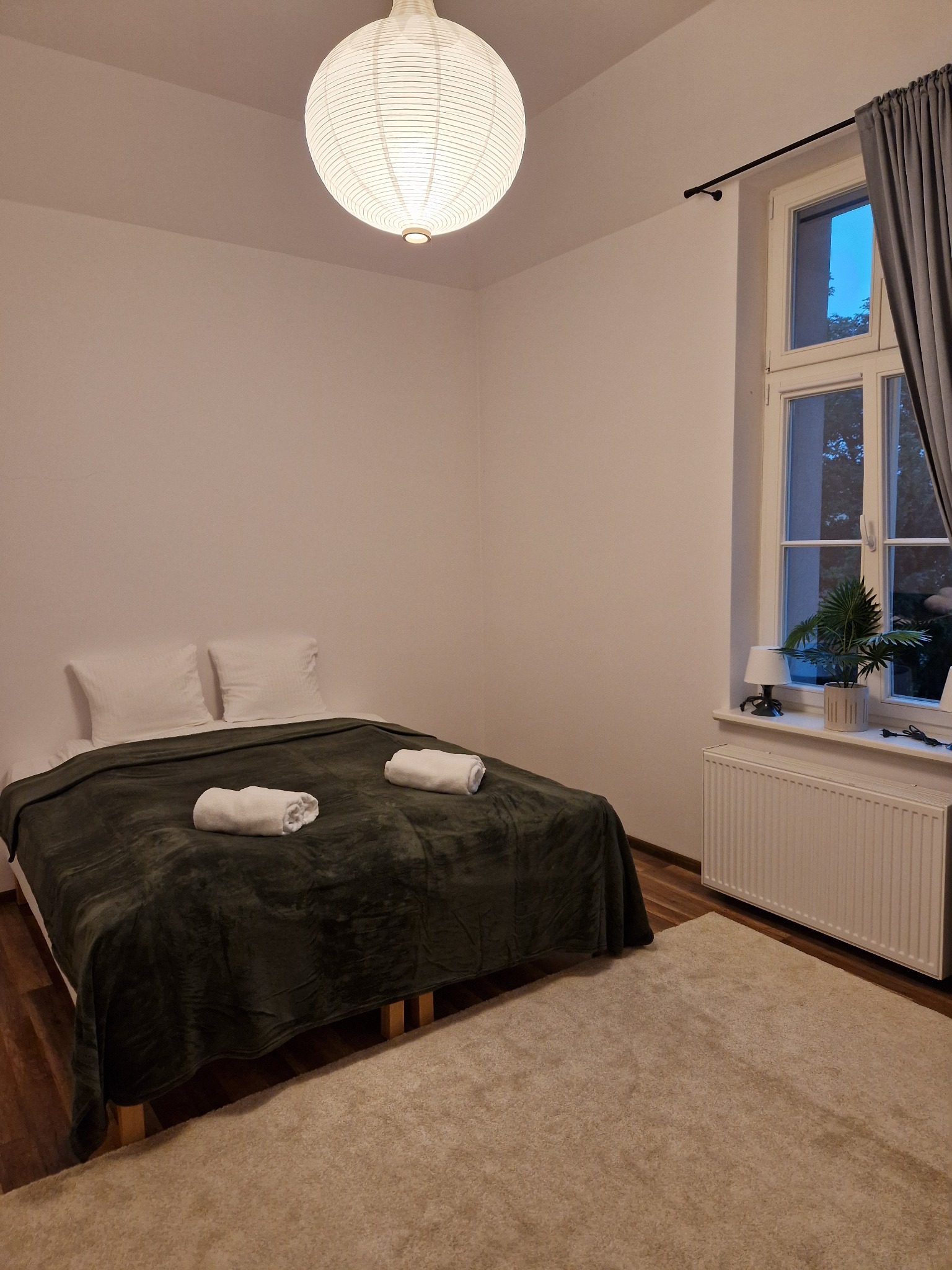 Przytulna sypialnia z łóżkiem z zielonym pledem i zwiniętymi ręcznikami, lampa w stylu japońskim, widok z okna na zewnątrz, dywan w kolorze ecru.