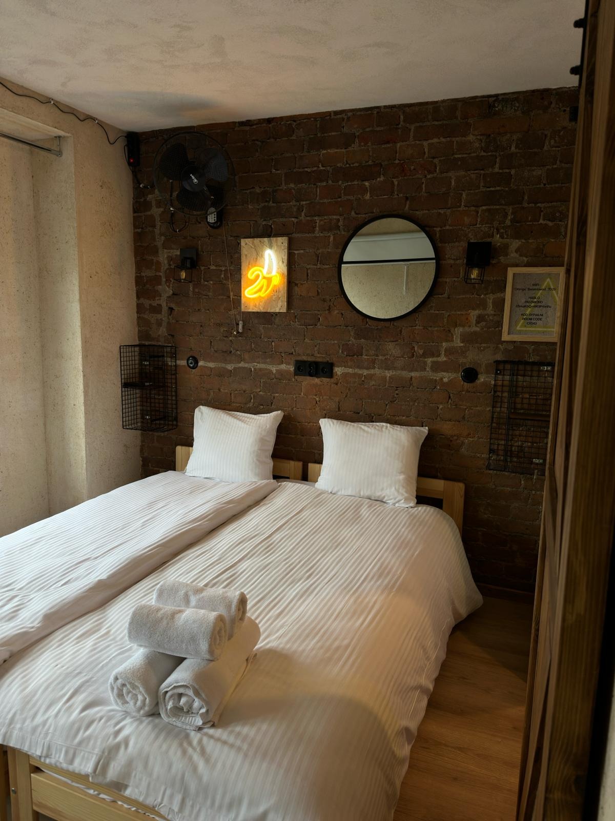 Pokój hotelowy z dwoma łóżkami pojedynczymi, stertą ręczników i ceglaną ścianą z neonem w kształcie banana.