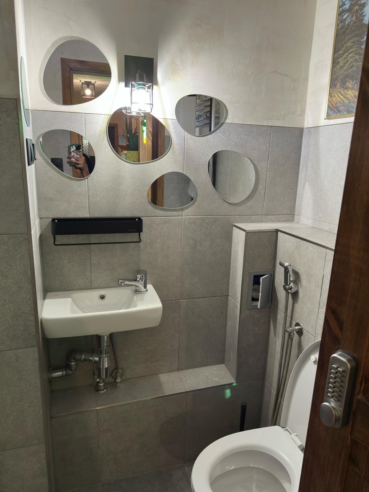 Nowoczesna łazienka z umywalką, toaletą i lustrami o nieregularnych kształtach na szarej ścianie wyłożonej płytkami.