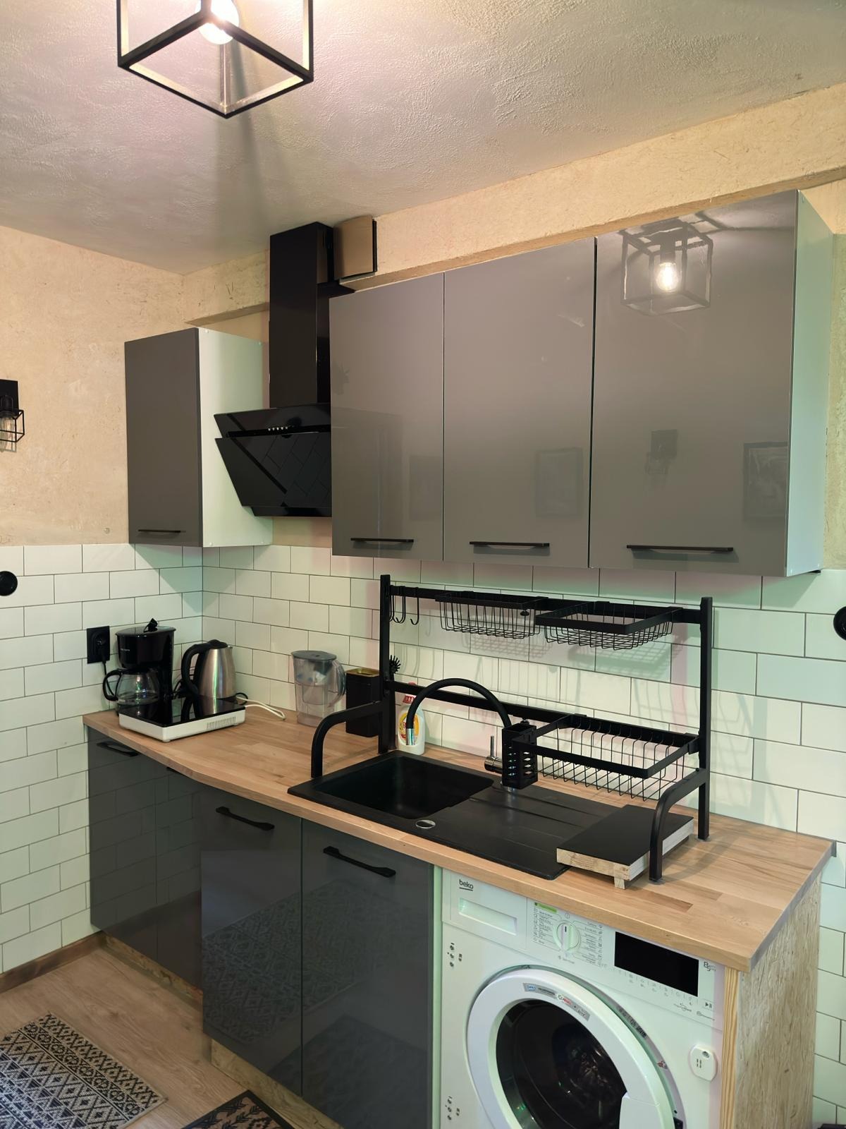 Kompaktowa kuchnia z szarymi szafkami na wysoki połysk, drewnianym blatem, czarnym zlewem i suszarką nad zlewem oraz pralką pod blatem. Widoczne białe płytki na ścianie i dekoracyjne lampy.