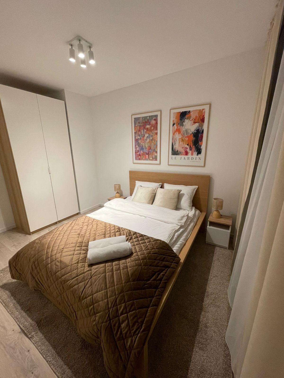 Ustawne wnętrze sypialni z łóżkiem o drewnianej ramie, brązową narzutą i białymi ręcznikami. Na ścianie wiszą dwa abstrakcyjne obrazy w ramkach.