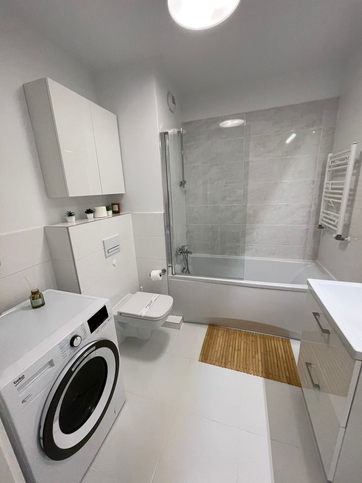 Jasna łazienka po remoncie: białe płytki, pralka Beko, wanna z parawanem, bambusowy dywanik i minimalistyczne dodatki. Czystość i nowoczesny design.