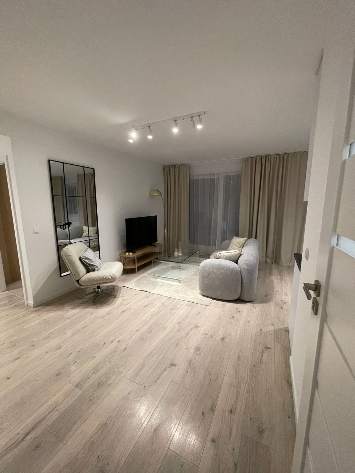 Jasne, nowocześnie urządzone wnętrze salonu z drewnianą podłogą, szarą sofą i fotelami. Widoczne okna z zasłonami i lustro na ścianie.