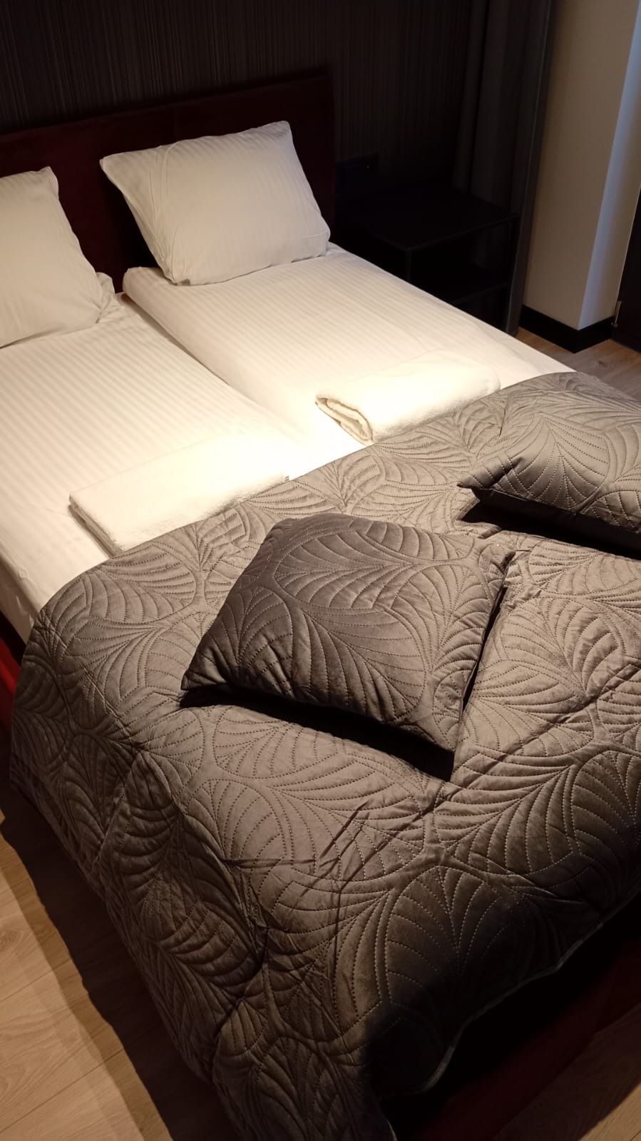Dwa łóżka pojedyncze z białą pościelą i szarą narzutą w liście, schludnie ułożone w pokoju hotelowym. Widoczne drewniane panele podłogowe i ciemne meble.