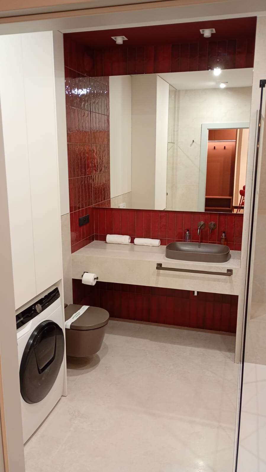 Nowoczesna łazienka z czerwonymi płytkami, umywalką w odcieniach brązu i pralką. Widoczne lustro i akcesoria łazienkowe. Minimalistyczny design.