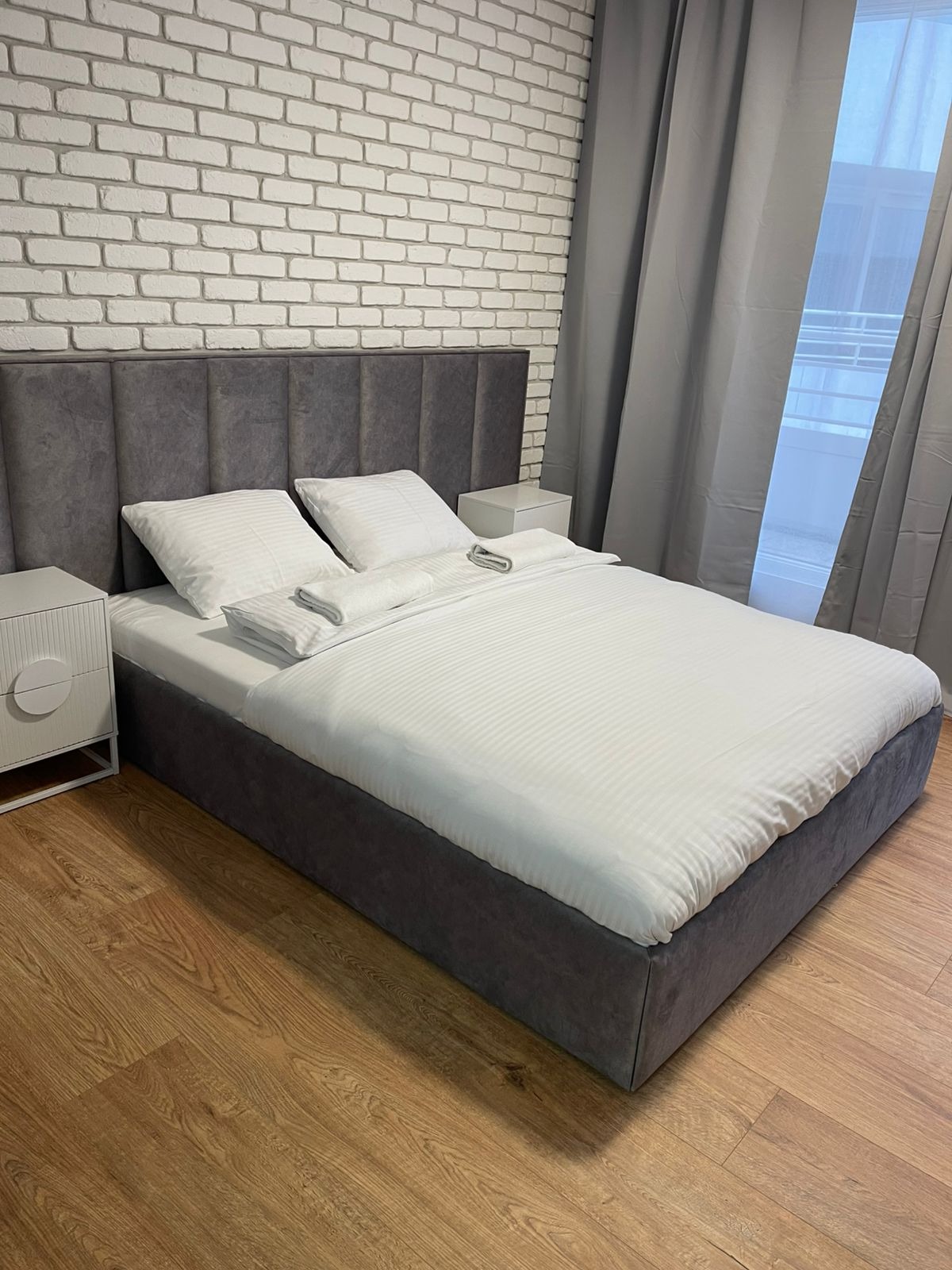 Czyste, białe prześcieradło na szarym łóżku z zagłówkiem, biała ceglana ściana w tle. Uprane tekstylia w pokoju hotelowym po usłudze sprzątania.