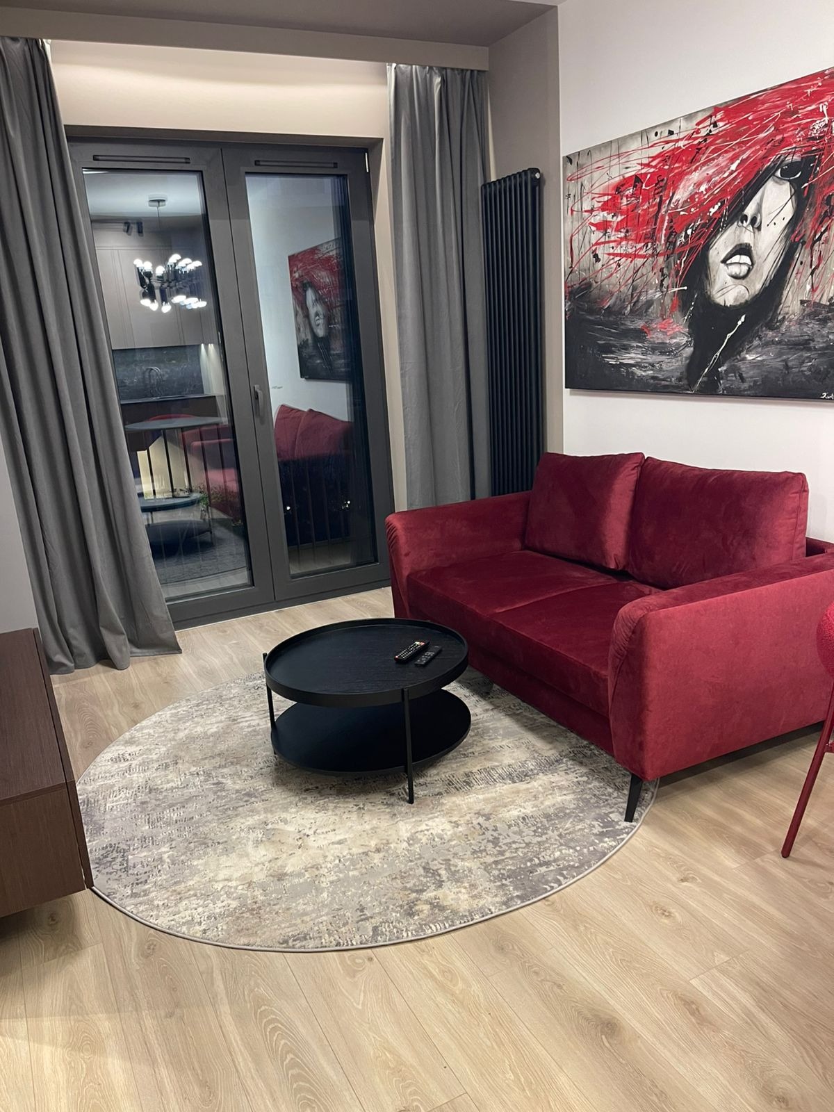 Wnętrze salonu po sprzątaniu: burgundowa sofa, okrągły dywan, czarny stolik kawowy z pilotami, obraz na ścianie, szare zasłony i parkiet. Widok z balkonu.