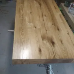 Blat stołu z jasnego drewna dębowego z wyraźnym usłojeniem i sękami, widoczny w warsztacie na tle betonowej podłogi i narzędzi.