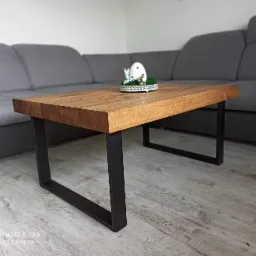 Stolik kawowy z blatem z jasnego drewna i czarnymi, metalowymi nogami w kształcie prostokątów, na blacie dekoracja wielkanocna z jajkiem i zajączkiem, w tle szara sofa.