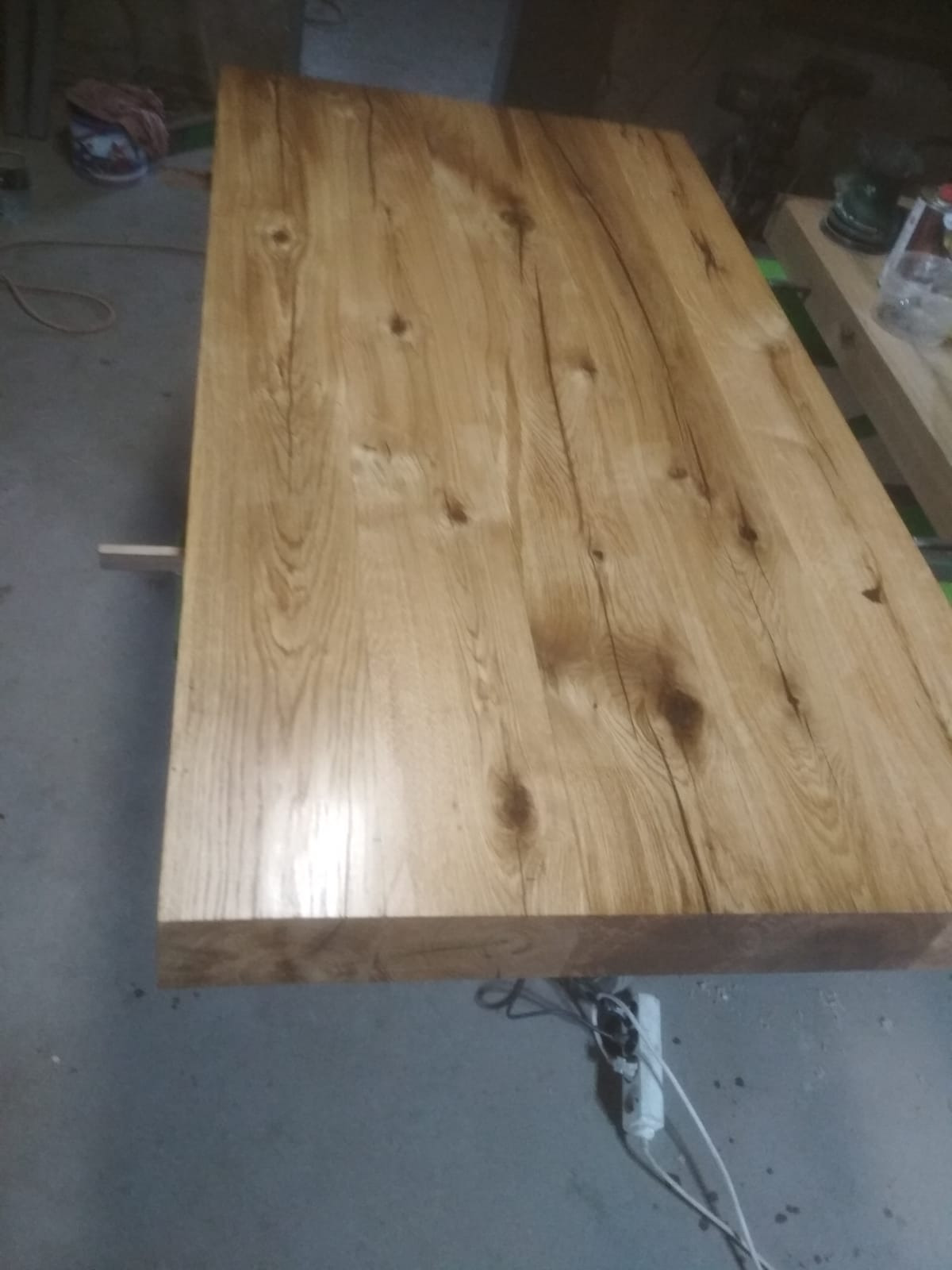 Blat stołu z jasnego drewna dębowego z wyraźnym usłojeniem i sękami, widoczny w warsztacie na tle betonowej podłogi i narzędzi.