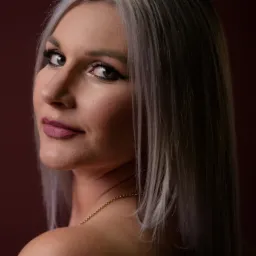 Portret kobiety z platynowymi włosami, spoglądającej przez ramię na tle bordowym. Delikatny makijaż, złoty łańcuszek na szyi. Ujęcie z bliska, z widocznym podpisem fotografa.