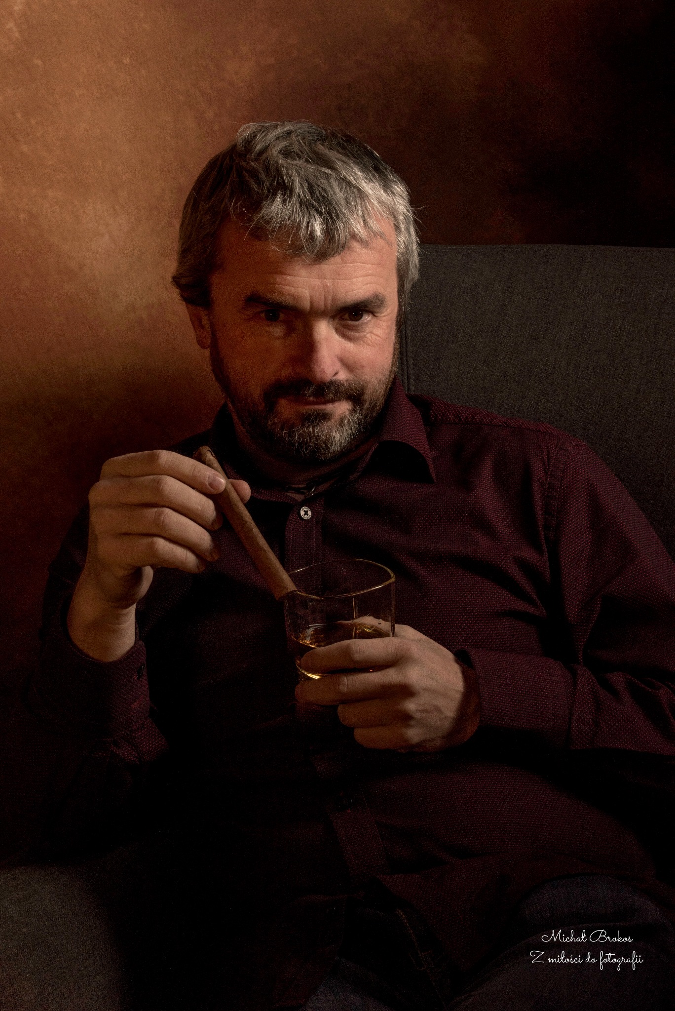 Mężczyzna w bordowej koszuli siedzi w fotelu, trzymając cygaro i szklankę whisky, zamyślony wzrok skierowany w stronę widza.