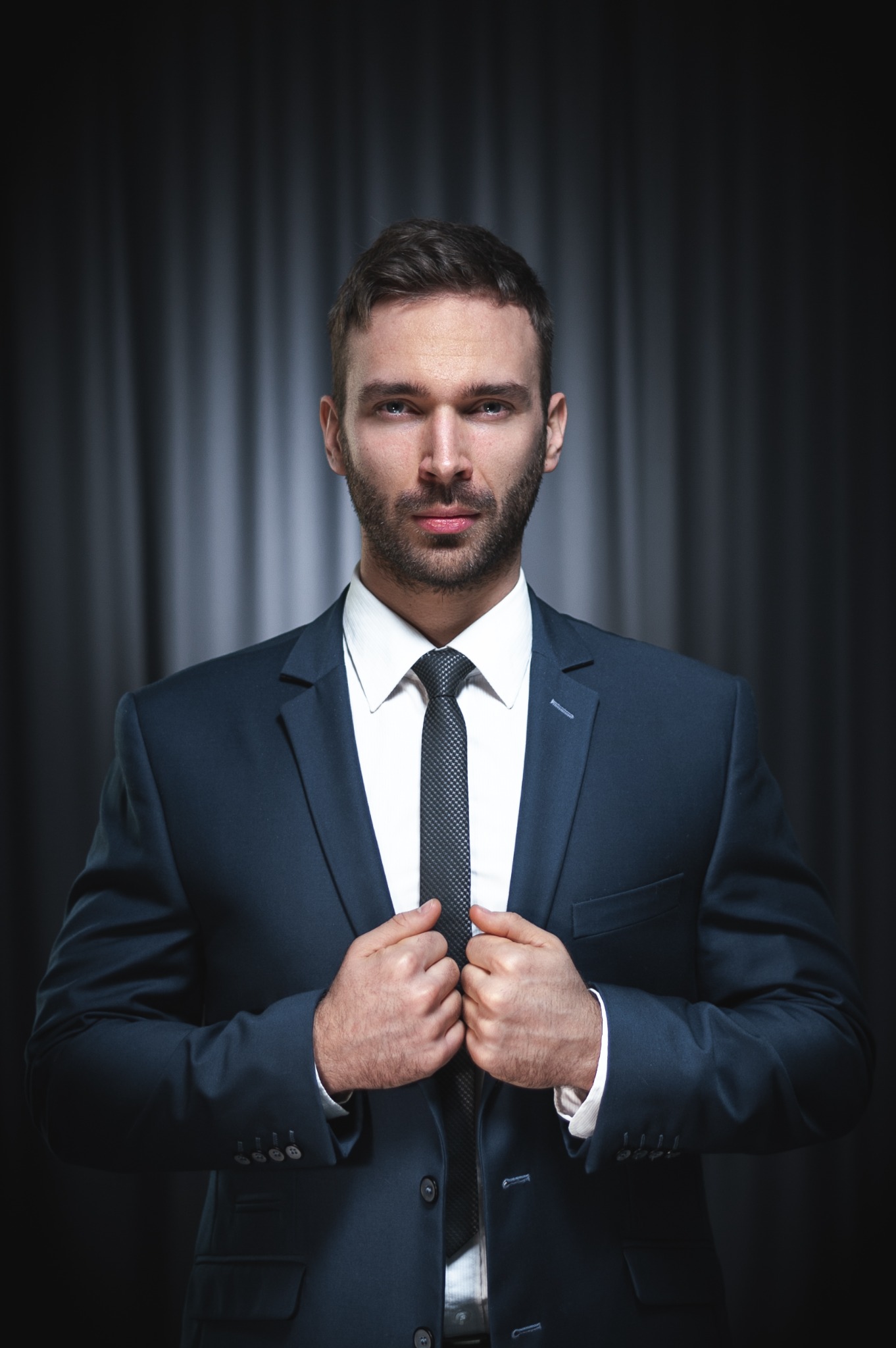 Mężczyzna w ciemnym garniturze i białej koszuli, poprawiający marynarkę na ciemnym tle z subtelnymi pionowymi fałdami.