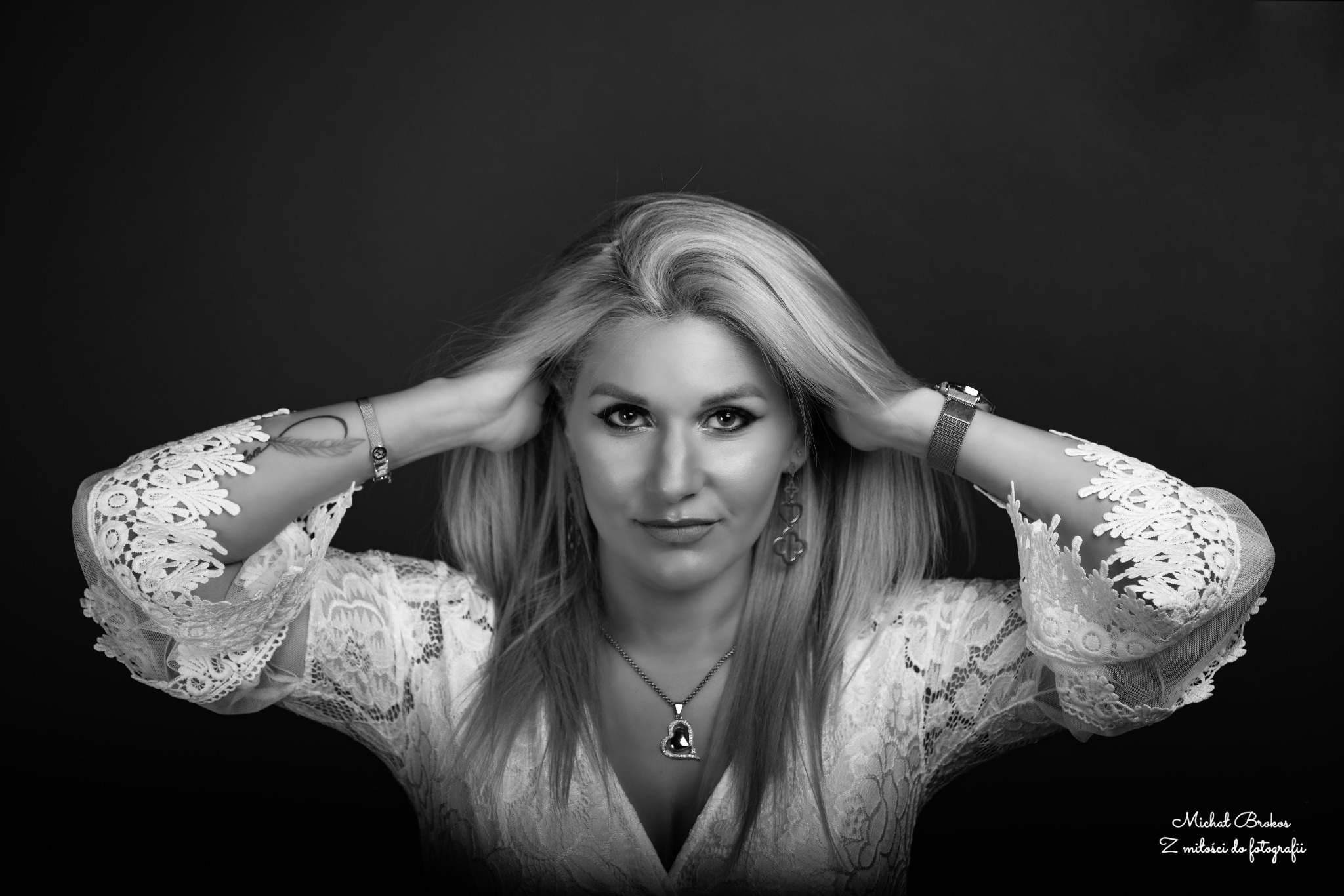 Portret kobiety w koronkowej bluzce, z blond włosami uniesionymi rękoma, na ciemnym tle, z widoczną biżuterią i tatuażem na ramieniu.