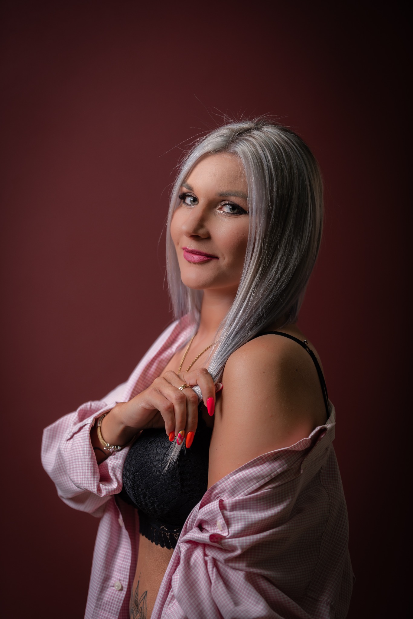Portret kobiety z platynowymi włosami, w rozpiętej koszuli i czarnym biustonoszu, pozującej na tle bordowej ściany, z widocznym tatuażem na brzuchu i neonowymi paznokciami.