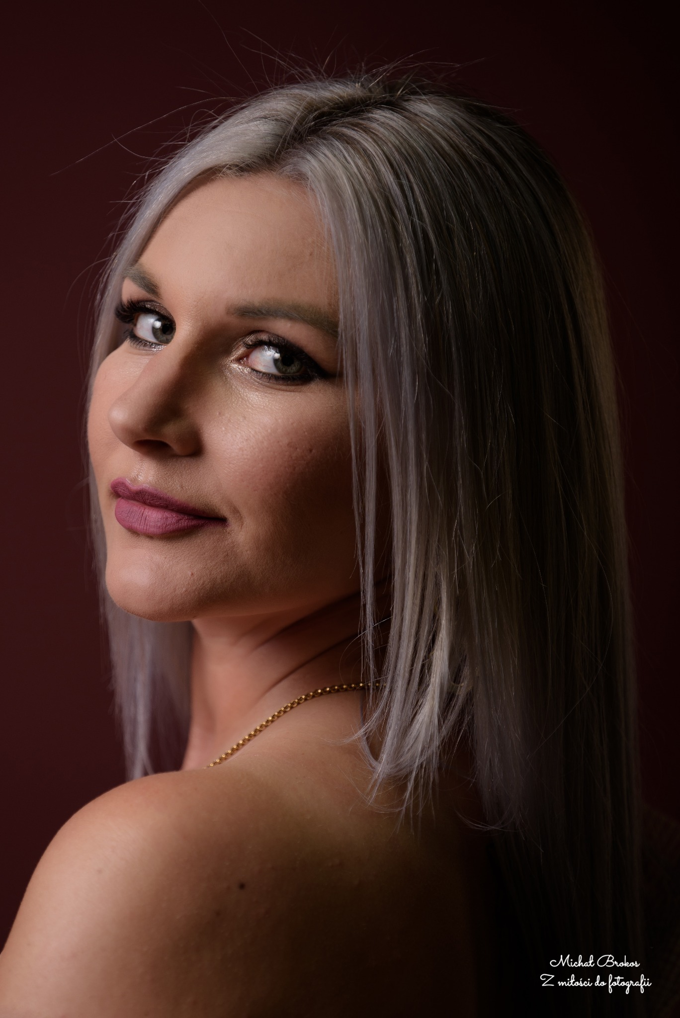 Portret kobiety z platynowymi włosami, spoglądającej przez ramię na tle bordowym. Delikatny makijaż, złoty łańcuszek na szyi. Ujęcie z bliska, z widocznym podpisem fotografa.