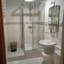 Wykończona łazienka z kabiną prysznicową, umywalką i toaletą w beżowej kolorystyce, z widocznym koszem na śmieci i ręcznikiem na podłodze.