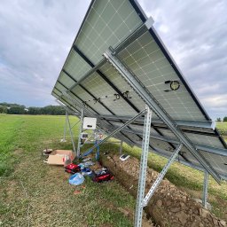 LDR Electrical Dariusz Karaś - Widok od tyłu na panele fotowoltaiczne w trakcie montażu na łące, widoczny wykop pod instalacje, narzędzia i skrzynki z akcesoriami. Na panelach widoczne żółte zabezpieczenia kabli.