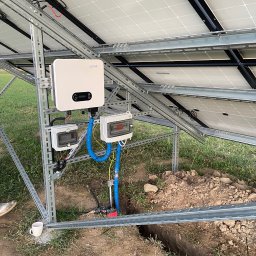 LDR Electrical Dariusz Karaś - Widok od spodu na panele fotowoltaiczne, inwerter Sofar i skrzynki elektryczne połączone niebieskimi peszlami, widoczny wykop w ziemi oraz kubek przy konstrukcji wsporczej.