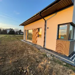 Nowoczesny dom jednorodzinny z beżową elewacją i brązowymi akcentami, widok z zewnątrz na tle zachodzącego słońca, teren wokół domu w trakcie prac ziemnych.