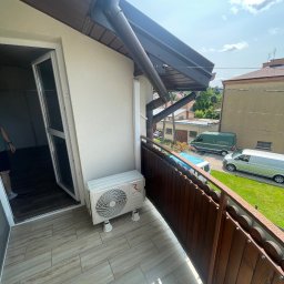 LDR Electrical Dariusz Karaś - Biała jednostka zewnętrzna klimatyzacji Rotenso na balkonie z drewnianą balustradą, widok na budynki i zaparkowane busy w słoneczny dzień.