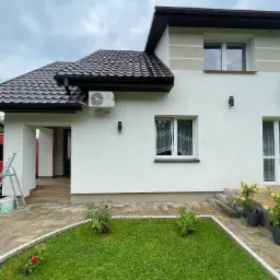 Biały dom z brązowym dachem i zamontowaną klimatyzacją na ścianie. Widoczny ogród z kostką brukową i roślinami. Drabina oparta o ścianę. Pochmurne niebo.