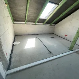 Surowy strych z oknem dachowym, instalacją elektryczną w peszlu i fragmentem listwy przypodłogowej. Widoczne bloczki betonowe i drewniana konstrukcja dachu.