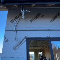 LDR Electrical Dariusz Karaś - Montaż instalacji elektrycznej na zewnątrz budynku w trakcie budowy. Widoczne kable i przewody przymocowane do ściany z membraną. W tle ekipa budowlana.