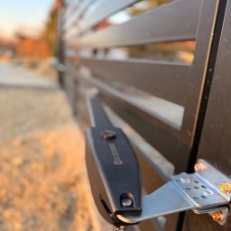 LDR Electrical Dariusz Karaś - Zamknięta, nowoczesna brama posesyjna z zamontowanym napędem elektrycznym, widoczne detale mocowania i przewód. Tło rozmyte, oświetlenie popołudniowe.